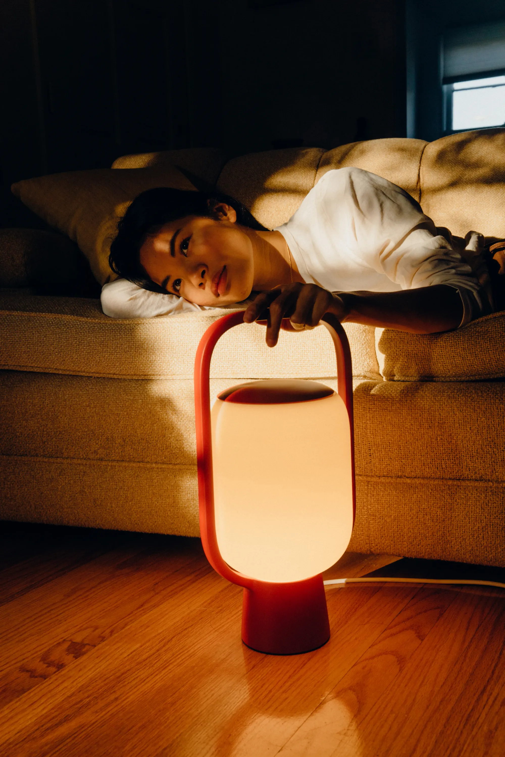 A woman lays on her couch, her face is partially lit by the Kero Table Light. She holds the handle of the lamp, which is firmly placed on the floor.