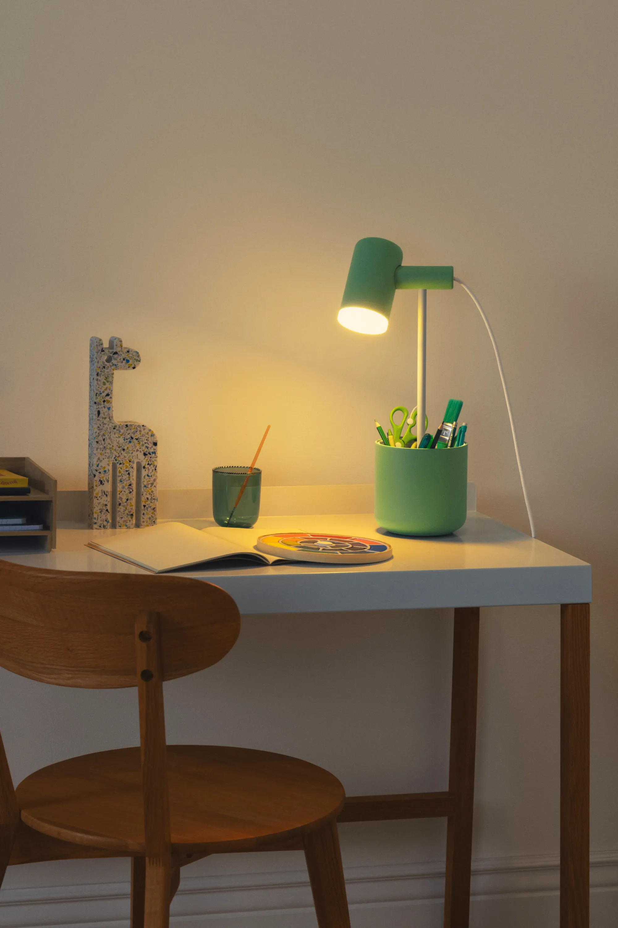 A table with watercolor materials is illuminated by Gantri’s Field Trip Table Light in sage green. The multi-purpose lamp also functions as a penholder for several writing materials and a pair of scissors.
