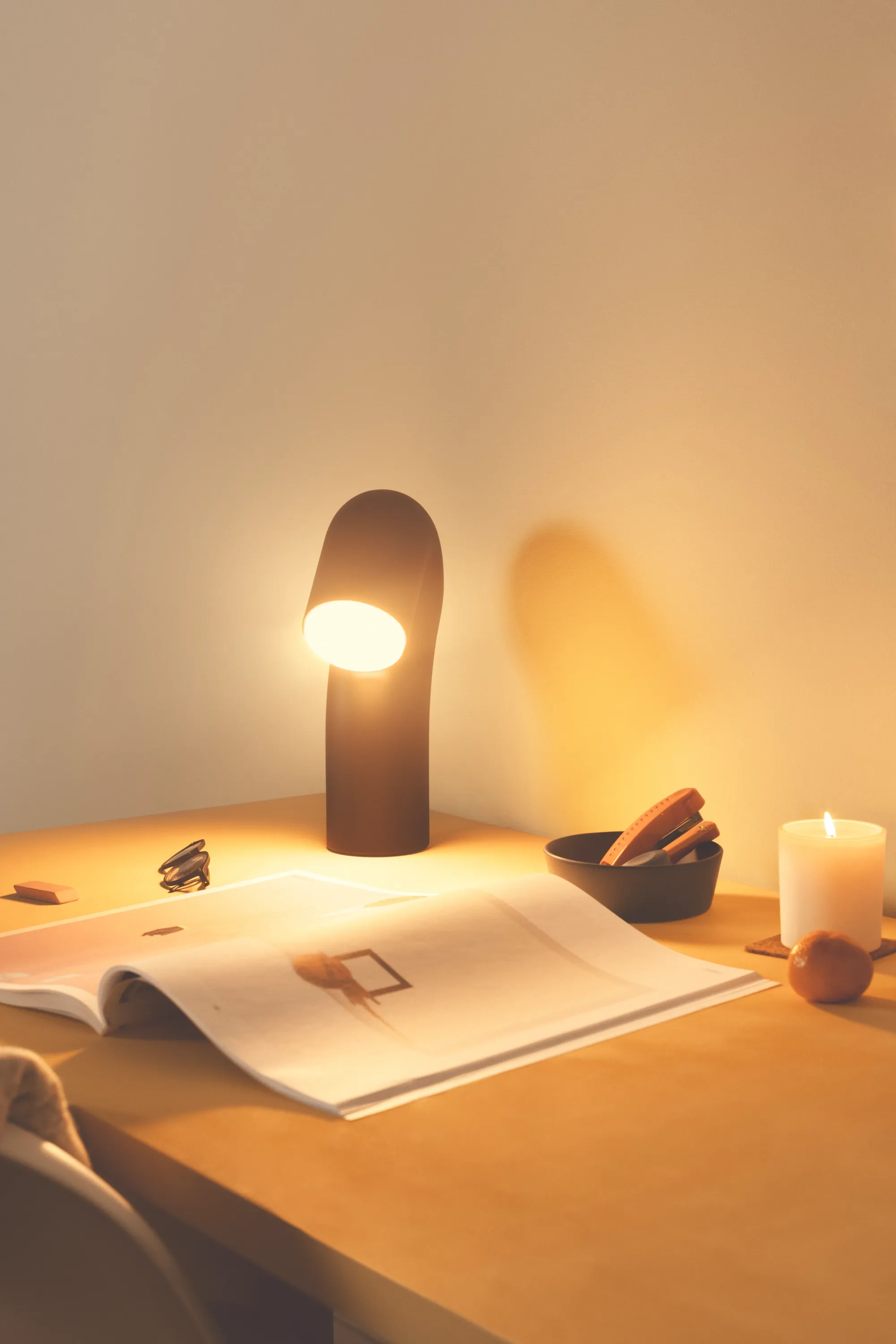 A home office table with an open magazine is being lighted by Gantri’s Smoothy table lamp and a small scented candle