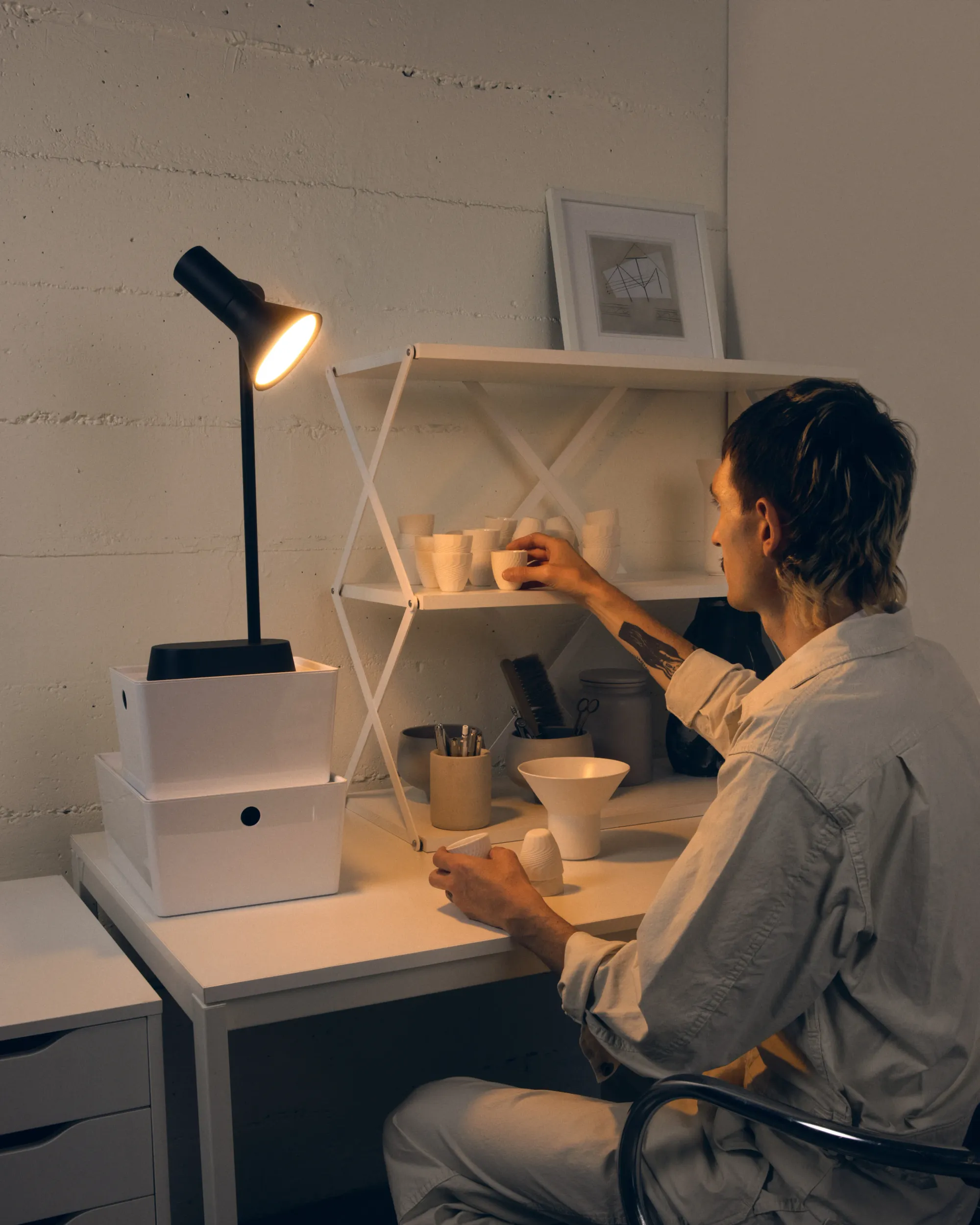 A man is placing small handmade ceramic mugs onto a shelf perched over his home office table. The area is illuminated by Gantri’s Gallery Task light table lamp.
