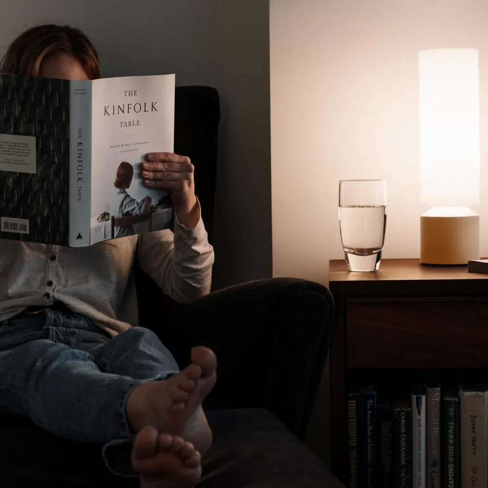A woman is sitting on a couch with her feet up while reading a book using light from the Lago table light perched on the table beside her