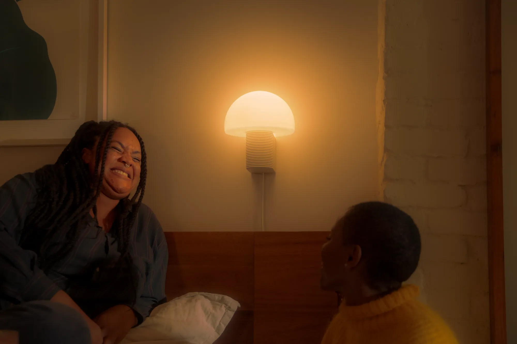 Two women are laughing together while hanging out. Their profiles are illuminated by light coming from a Gantri Arintzea Wall Light emitting warm light. 