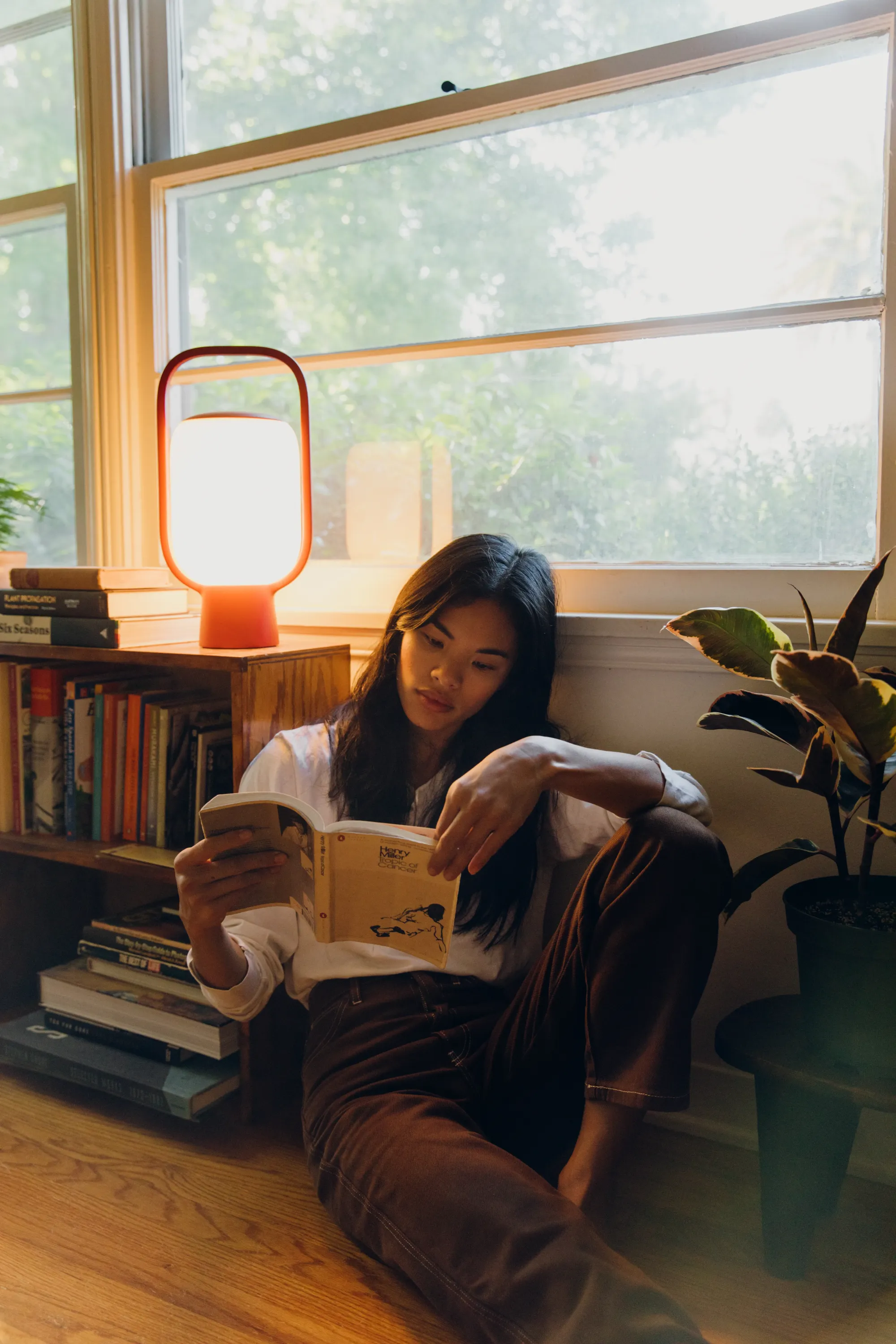 A woman is reading by her window and small wooden bookcase filled with books. Natural light and light from Gantri’s Kero Table Light helps her read. 