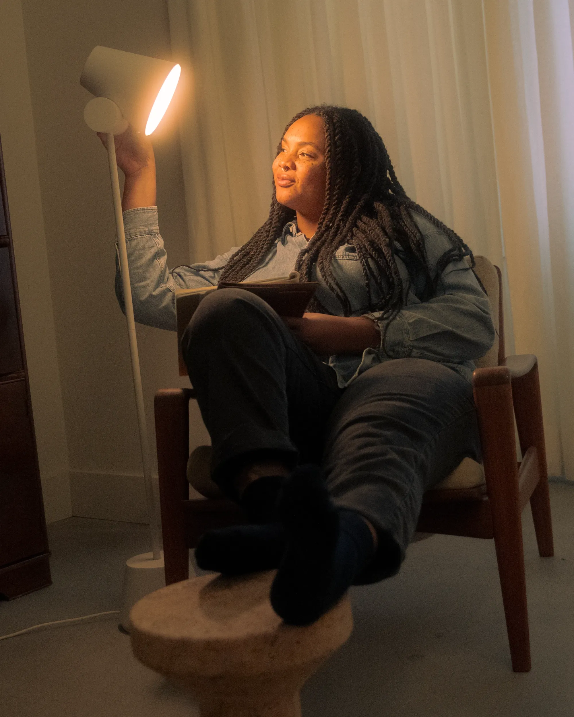 A woman sits on her living room chair. She is holding a tablet and a thin magazine in one hand,  and touching Gantri’s Aim Floor Light with the other. The light illuminates her face and frame